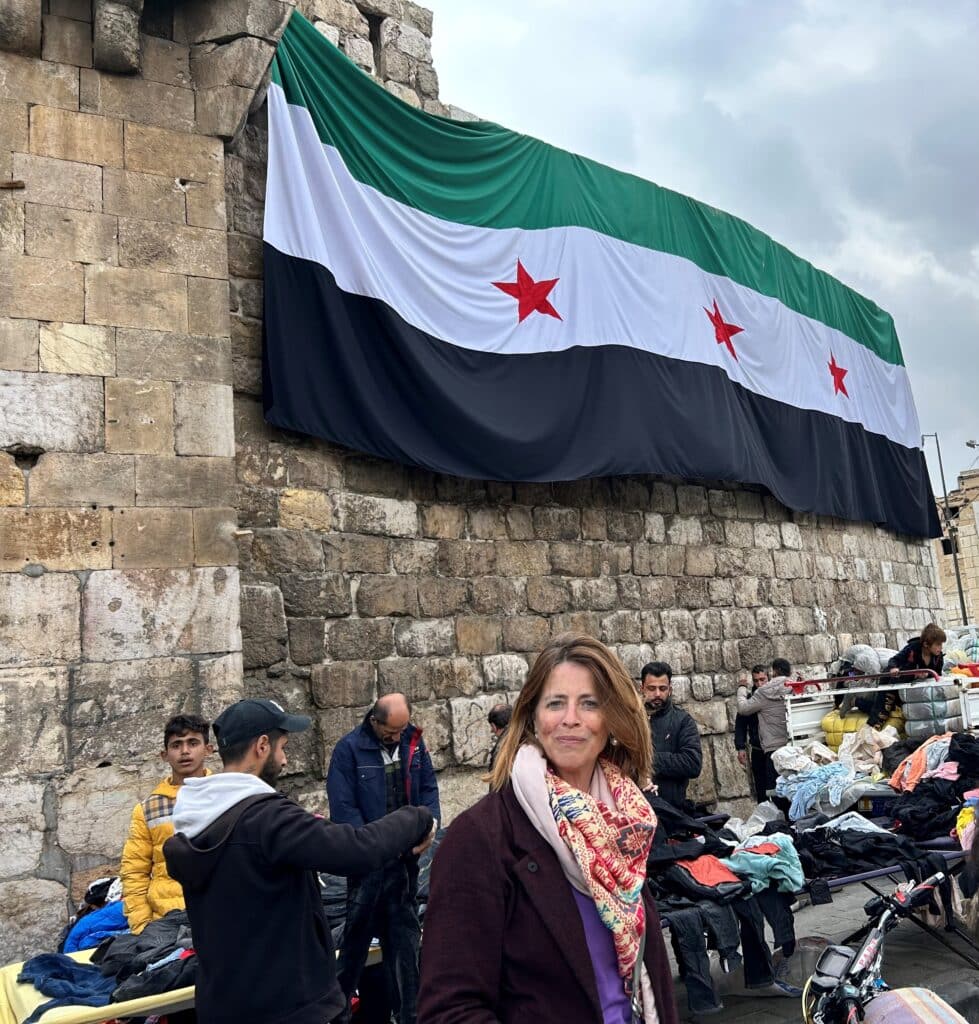 Petra Stienen tijdens haar eerste bezoek aan Syrië na de val van het Assad regime, vlag wappert, mannen in de achtergrond staan achter tafels met daarop hopen kleding.