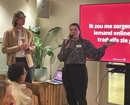 Twee sprekers tijdens de HVN-avond over tradwives en de manosphere. Een van hen spreekt met een microfoon en gebaart naar het publiek, terwijl de ander naast haar staat. Op de achtergrond is een presentatie met een rode dia te zien.